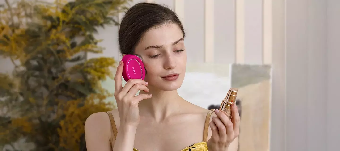 A young woman with sleek dark hair in yellow dress using FOREO BEAR 2 microcurrent device and conductive SUPERCHARGED serum 